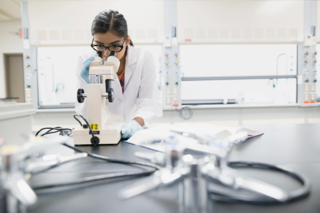 female scientist in lab space with windows looking through a microscope