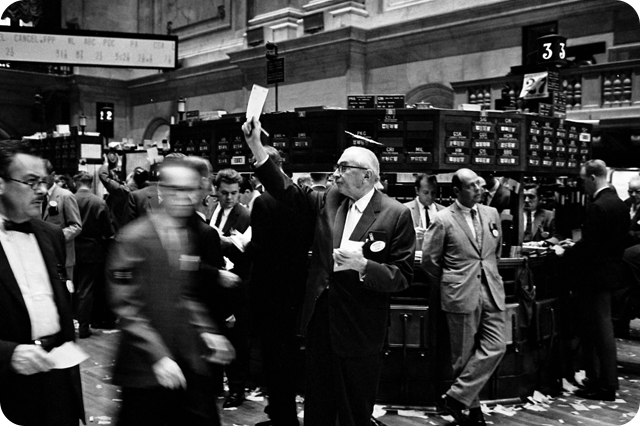 Black and white photo of a busy stock exchange floor with men in suits, one prominently holding up a paper