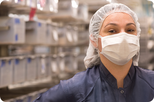 A smiling woman, wearing a hairnet and a face mask, stands in a manufacturing facility.
