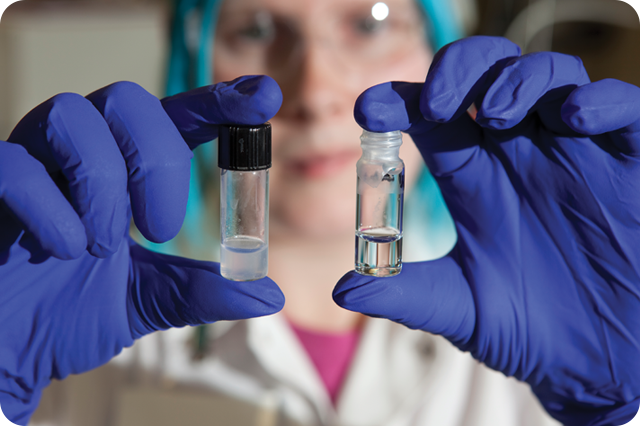 Close-up of a scientist's hands, wearing blue nitrile gloves, holding up two small vials containing clear liquids.