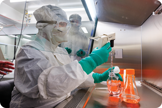 A scientist in full protective gear working in a laboratory, using a pipette to transfer liquid into a flask inside a sterile environment.